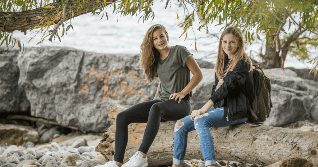 image - California Mental Health Two smiling teens sitting outdoors on a log, fostering growth through industry and overcoming inferiority, building confidence.