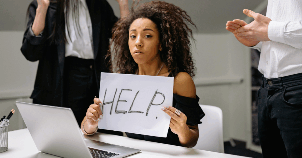 image - California Mental Health Sadistic Personality Disorder: Woman at desk holding 'Help' sign, colleagues nearby, depicting workplace stress or abuse.
