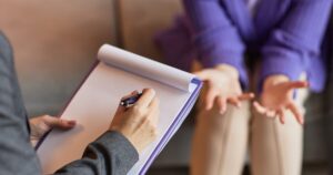 - California Mental Health Therapist takes notes on a clipboard while a client gestures with open hands during a conversation.