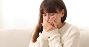 - California Mental Health Woman with brown hair in a cream sweater covers her mouth with both hands, eyes closed, showing distress or sadness at home.