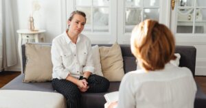 - California Mental Health Two women sit on a gray sofa in a bright living room, talking in a counseling session.