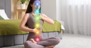 - California Mental Health Woman sits cross-legged on a carpet in a bedroom, meditating with colorful chakra lights along her chest, throat, abdomen and pelvis area.