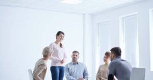 - California Mental Health Woman stands and speaks to a seated, diverse team in a bright office meeting room.