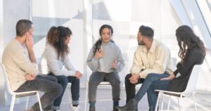 - California Mental Health Five people sit in a circle in a bright, modern space; a woman in a gray sweater speaks with hand gestures while others listen.