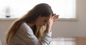 - California Mental Health A woman sits with her head bowed and hands clasped against her forehead, appearing stressed or focused in a bright room.