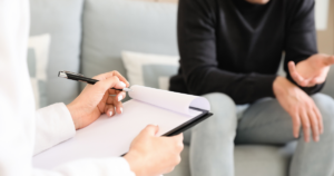 - California Mental Health Person writes on a clipboard with a pen during a counseling or interview session while a patient sits on a sofa and gestures with their hands in the background