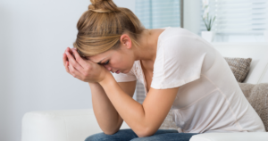 - California Mental Health Woman sitting on a white couch, head bowed, hands covering her face, looking distressed or overwhelmed.