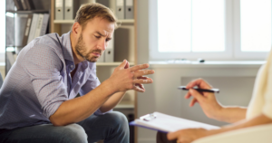 - California Mental Health Man in a counseling session, leaning forward with hands clasped, looking worried as a clinician takes notes on a clipboard.