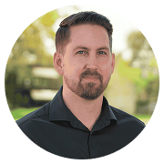 Portrait of a man with short dark hair and a trimmed beard, wearing a black collared shirt, outdoors with greenery in the background.