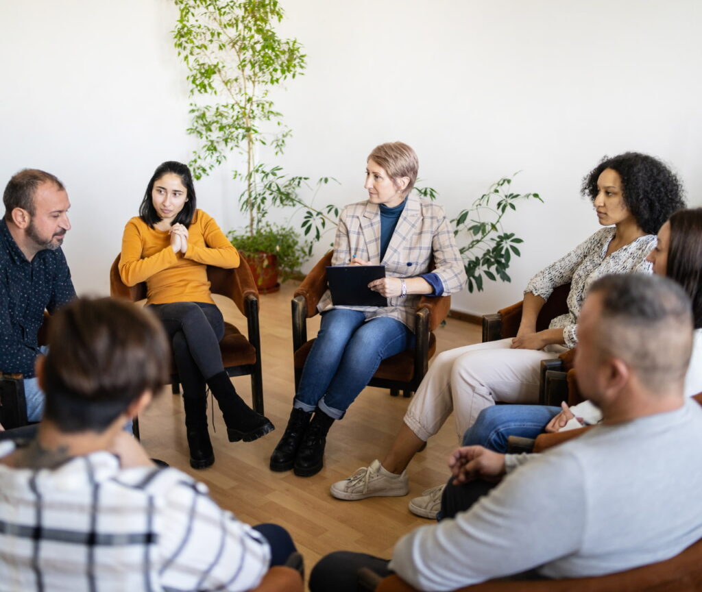 Group of adults seated in a circle in a bright room with several potted plants; facilitator holds a clipboard and leads the discussion.
