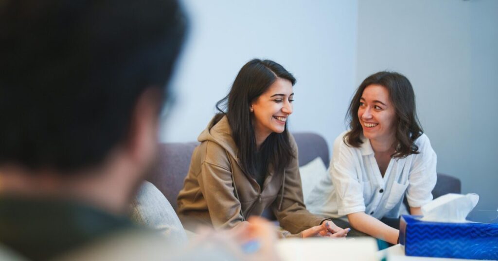 Two women sit on a sofa, smiling and talking, with a blurred person in the foreground suggesting a small group discussion in a living room.