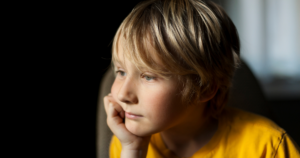 Young boy with blond hair in a yellow shirt rests his chin on his hand, gazing pensively to the left.