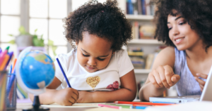 Young girl focused on drawing at a desk with a blue pencil, globe and colored pencils nearby; an adult assists in a bright learning space.