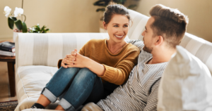 A smiling couple sits on a light sofa, leaning toward each other as they chat in a cozy living room.