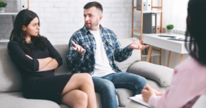 Man on a sofa speaks with animated hand gestures as a woman listens with folded arms; therapist sits nearby taking notes in a bright office.