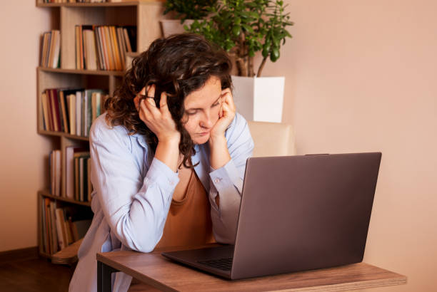 Woman resting her head in her hands while looking at a laptop on a wooden desk in a cozy home office.